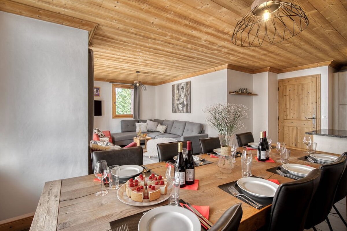 A cozy chalet-style open-plan living and dining space with a long wooden table set with cake and wine, leading to a comfy sofa area.