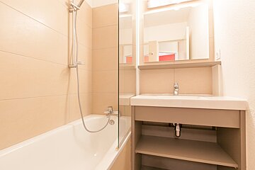 A modern bathroom features beige tiled walls, a white bathtub with shower, and a sink vanity with a mirror reflecting a red door.