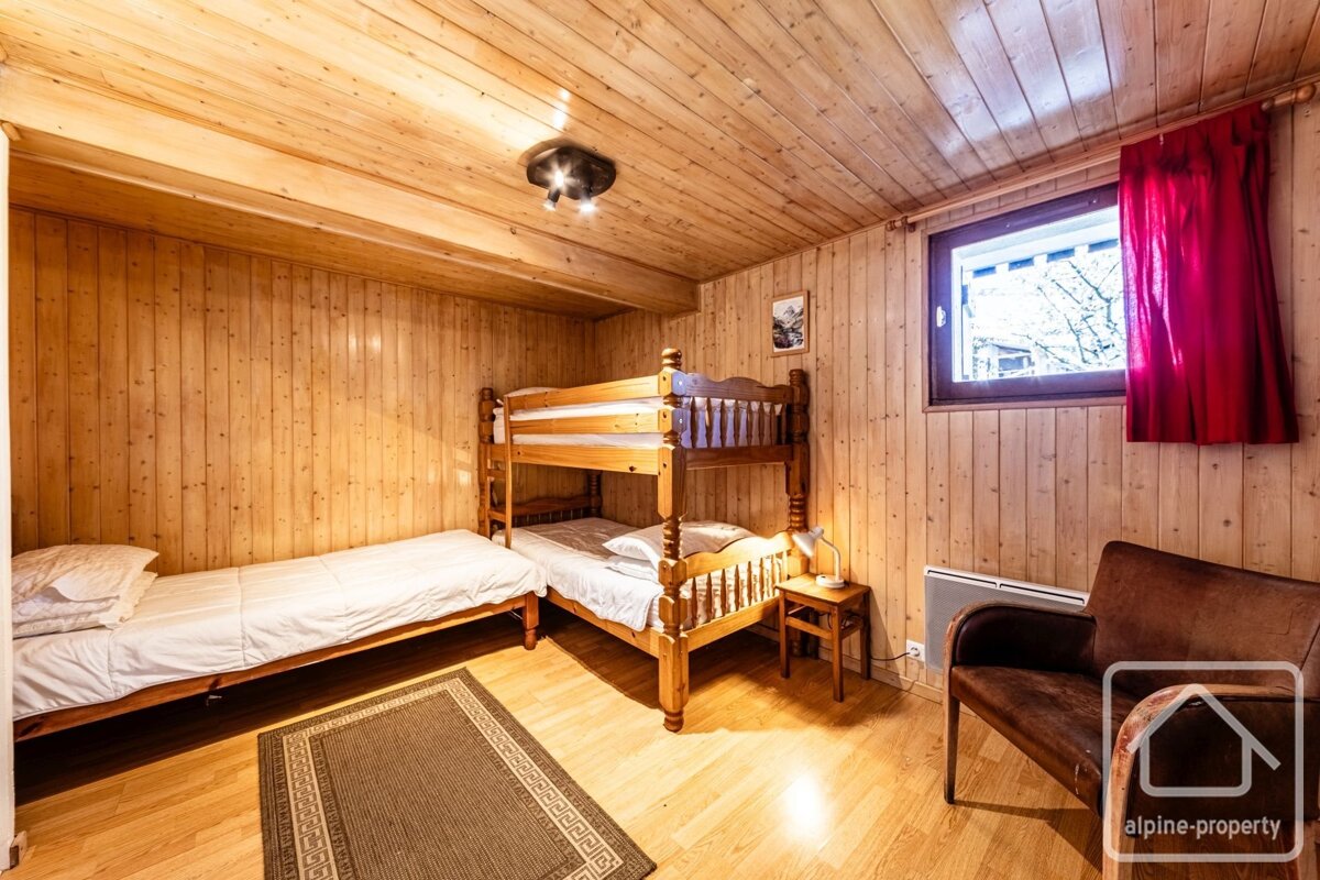 Rustic wood-paneled bedroom featuring bunk beds, a single bed, armchair, and a window with red curtains.