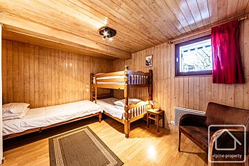 Rustic wood-paneled bedroom featuring bunk beds, a single bed, armchair, and a window with red curtains.