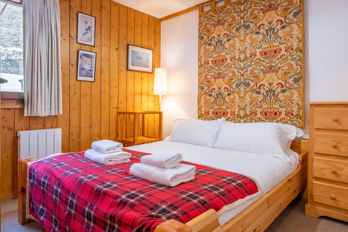 A cozy bedroom features wood-paneled walls, a bed with a red plaid blanket, white bedding, and a vibrant tapestry. Snow is visible outside the window.