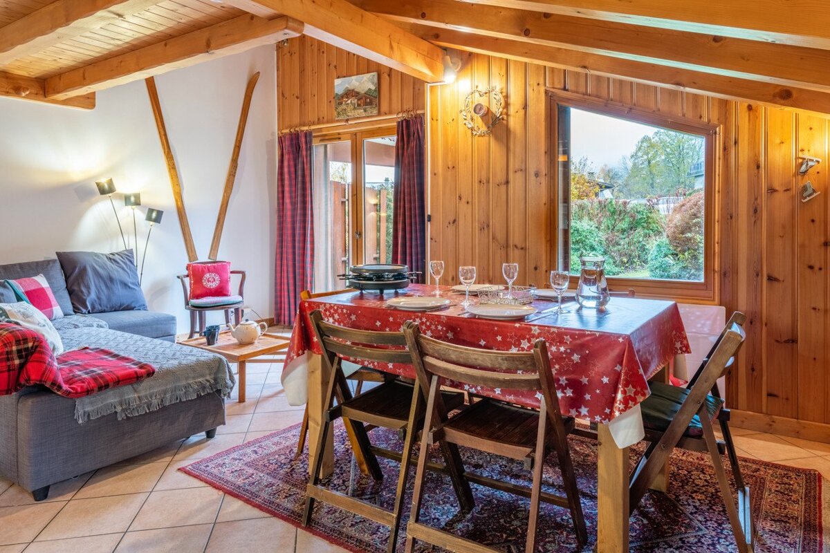 A rustic, wood-paneled chalet interior featuring a dining table set with a raclette grill, a comfy sofa, and a window looking outdoors.