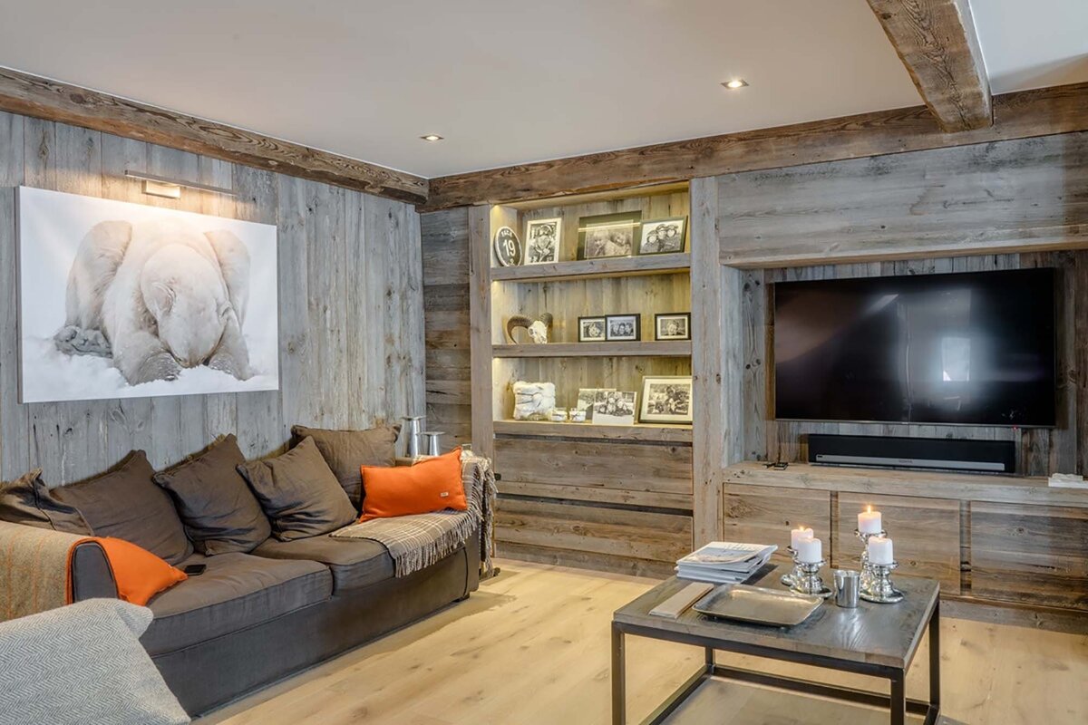 A cozy, rustic living room featuring reclaimed wood walls, a dark sofa with orange accents, polar bear art, built-in shelves, a TV, and a coffee table with candles.
