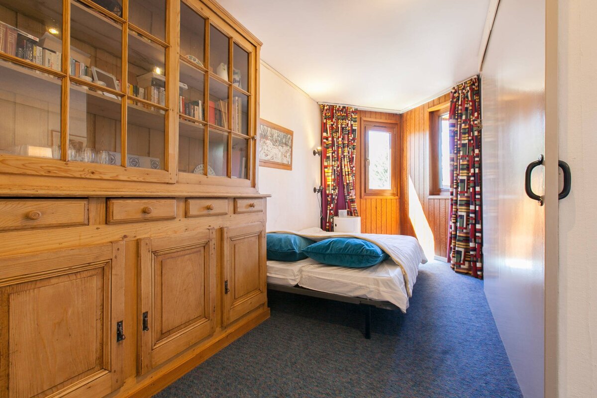 Cozy bedroom with a large wooden display hutch, a bed with blue pillows, blue carpet, and windows with colorful curtains and wood paneling.