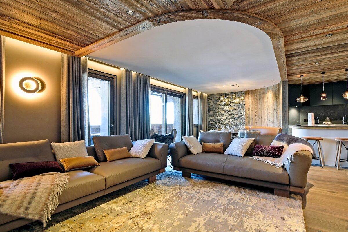 Modern chalet living room featuring warm wood ceilings, two taupe sofas with pillows, a textured rug, stone wall, and open kitchen with bar seating.