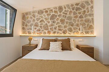 A bedroom with a prominent rustic stone accent wall above the bed, illuminated by warm backlighting and modern pendant lights. Neutral bedding and wood nightstands.