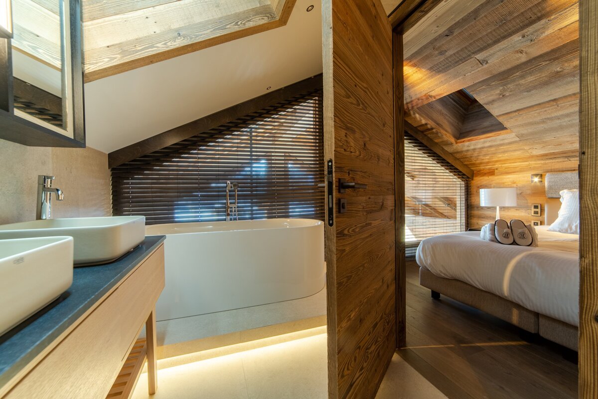 Cozy wooden chalet interior featuring a bathroom with a sleek bathtub, looking through an open door into a warm, rustic bedroom.