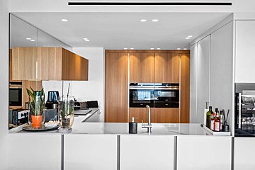A clean, modern kitchen with a white island, light wood cabinetry, integrated ovens, and a large mirrored wall reflecting kitchen elements.