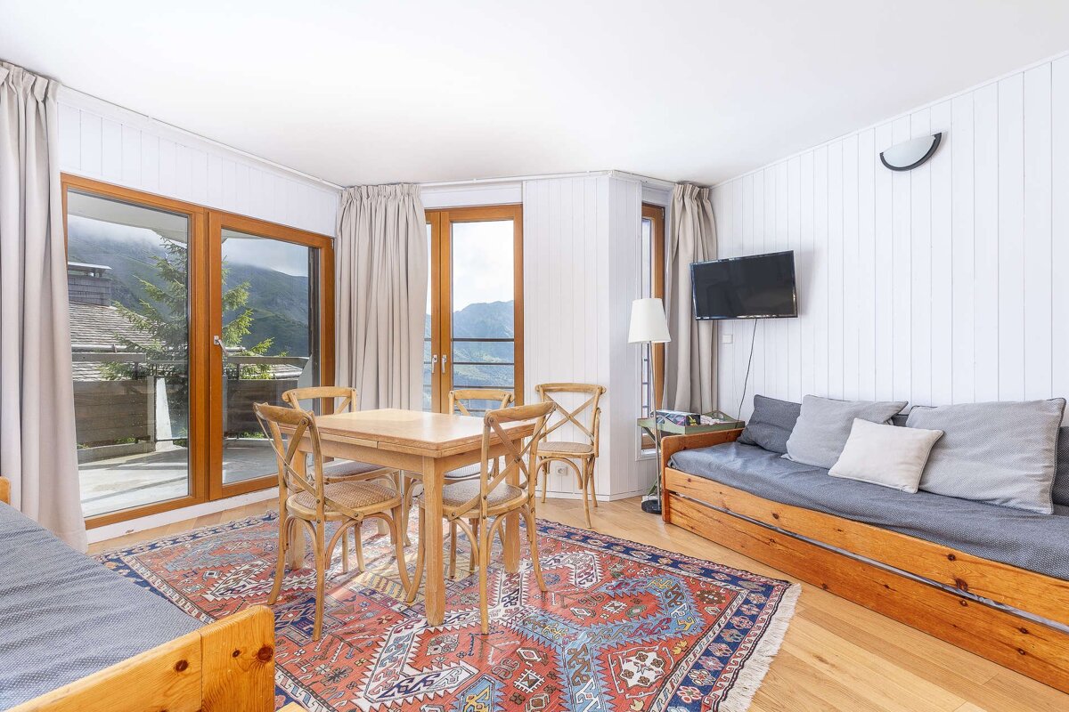 Bright, cozy room with mountain views, featuring a wooden dining table, daybed with cushions, wall-mounted TV, hardwood floor, and a colorful rug.
