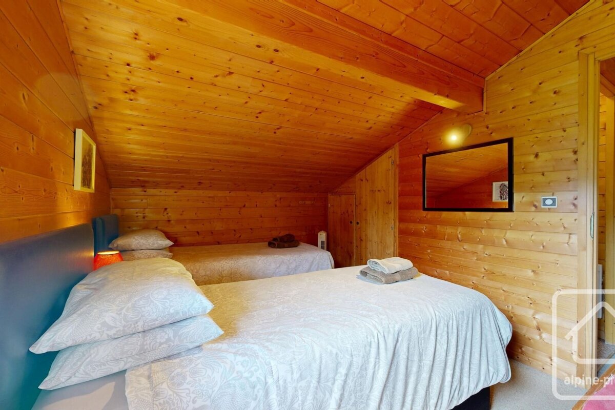 A cozy, rustic bedroom with sloped wood-paneled walls and ceiling, featuring two twin beds with white bedding, and a mirror.