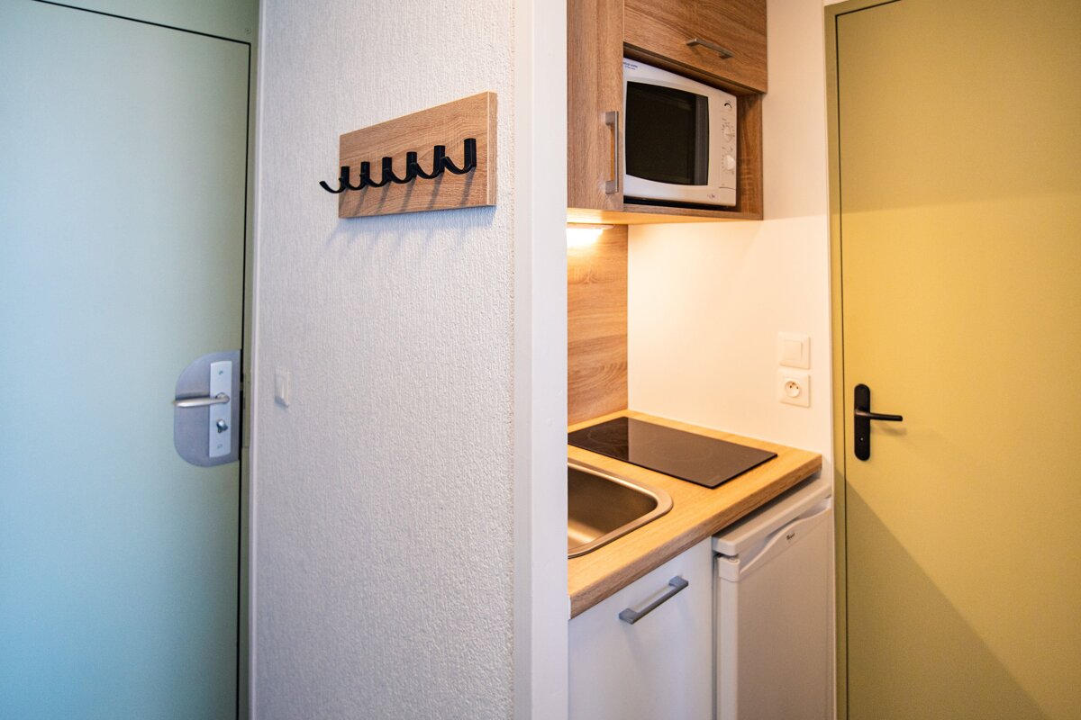 A compact kitchenette with microwave, sink, and stovetop, flanked by a coat rack on one wall and two doors in different colors.