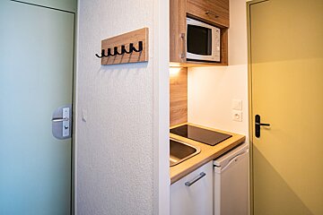 A compact kitchenette with microwave, sink, and stovetop, flanked by a coat rack on one wall and two doors in different colors.