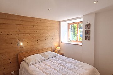 A cozy bedroom with a bed, white bedding, and a wooden headboard against a wood-paneled accent wall. A window provides natural light.