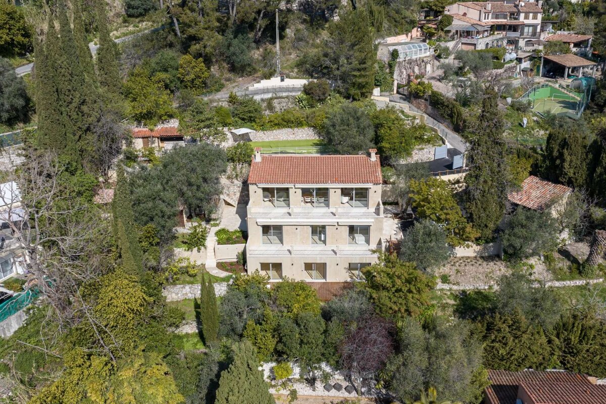 Aerial view of a large house with a red tile roof & balconies, surrounded by dense trees on a hillside. Other homes & a tennis court are visible.
