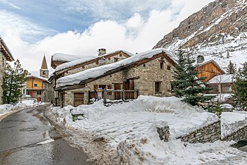 Charming snowy village street with stone houses, a prominent church spire, and a towering, snow-dusted mountain backdrop.