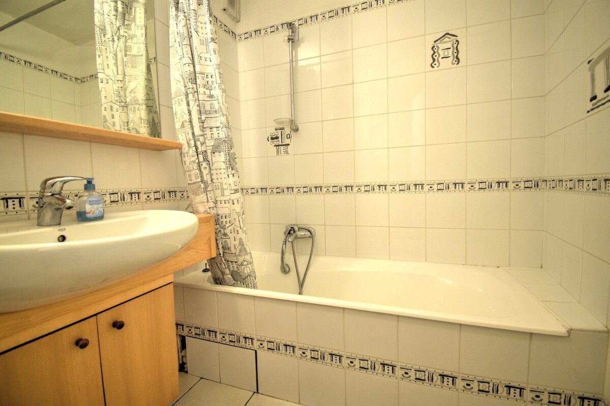 A white-tiled bathroom with a bathtub, sink, wooden vanity, and a cityscape-patterned shower curtain reflected in a mirror.