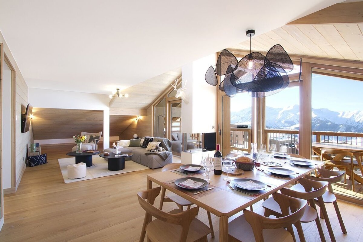 Modern chalet interior with open-plan living and dining areas, featuring a cozy sofa, stylish table, and panoramic snowy mountain views from large windows.