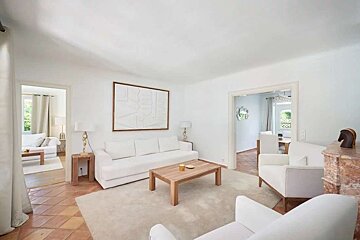 A clean, bright white living room featuring a long white sofa, wooden coffee table, abstract wall art, and terracotta-tiled floors.