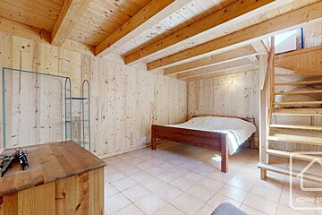 A cozy room with light wood paneling, exposed beams, and a tiled floor. It features a wooden bed, dresser, metal rack, and a prominent wooden staircase.