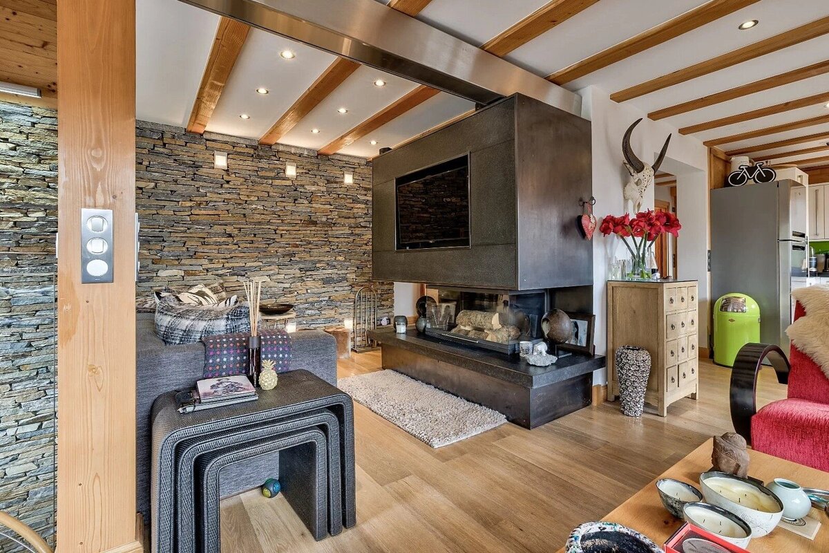 A cozy, modern living room with a stone accent wall, wood-beamed ceiling, and a sleek fireplace with a TV mounted above, creating a warm, inviting ambiance.