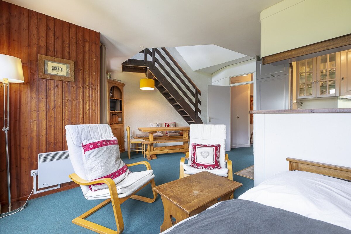 A rustic, cozy apartment interior with wood-paneled walls, armchairs, a wooden dining table, a visible staircase, and a bed in the foreground. Teal carpet throughout.