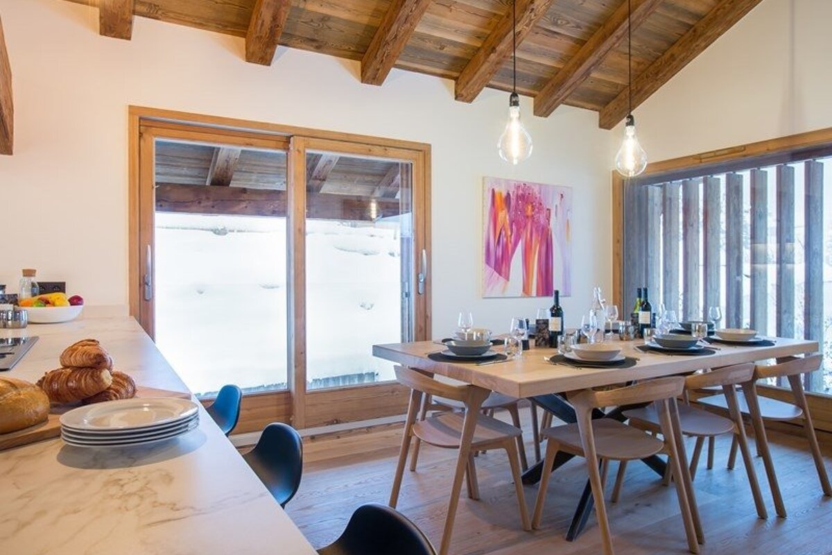 Cozy chalet dining room featuring a set wooden table, hanging bulb lights, and glass doors looking out onto a snowy landscape.