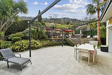A large, tiled outdoor patio with a dining set and lounge chair, leading to a lush garden with red umbrellas, a wooden deck, and distant mountains.