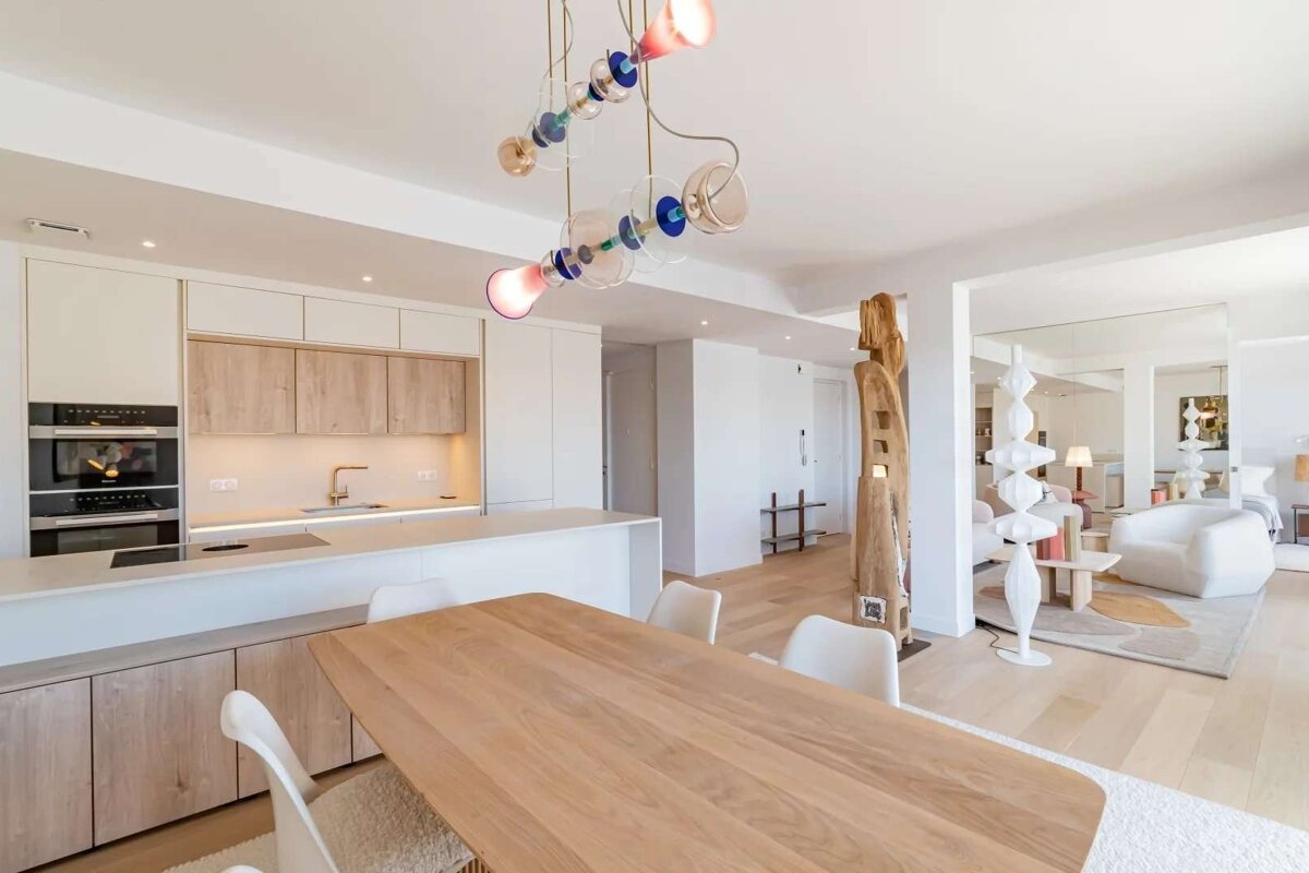 Bright, open-plan kitchen and dining space with light wood, white cabinetry, and a unique, colorful chandelier. Contemporary furnishings and art.