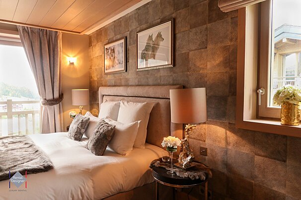 A luxurious bedroom features a large bed with faux fur, wolf artwork on a stone-textured wall, and warm lighting. A balcony offers a view of nature.