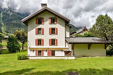 A multi-story white house with orange shutters stands on a green lawn against a backdrop of cloudy mountains.