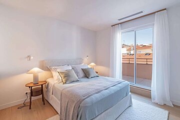 A bright, modern bedroom with a comfortable bed, nightstands, and soft lighting. A large glass door opens to a balcony overlooking a cityscape with red roofs.