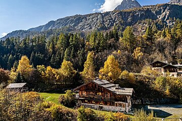 A wooden chalet nestled in a vibrant autumn forest with a majestic mountain backdrop under a clear blue sky.
