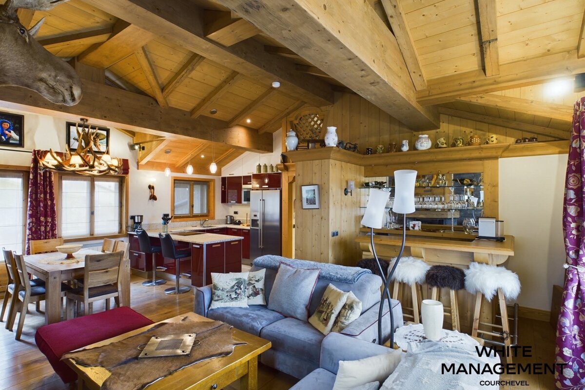 A rustic-chic chalet interior with wood beams, featuring a cozy living room, dining area, modern kitchen, and a bar. A moose head adds character.