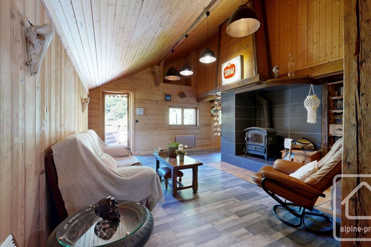 A rustic wood-paneled living room with a large fireplace, wood stove, comfy seating, and a Stella Artois sign, exuding a cozy lodge atmosphere.