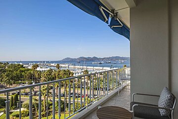 Sunny balcony view of a bustling coastal marina with boats, blue sea, palm trees, distant mountains. Balcony has table, chair, blue awning.
