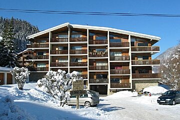 A large, multi-story wooden chalet with many balconies stands in a snowy mountain resort. Cars are parked in front, surrounded by snow-covered trees under a blue sky.