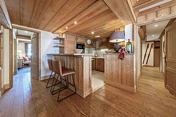 Cozy, rustic kitchen and hallway in a wooden chalet. Features wood-paneled walls, ceiling, floor, kitchen with bar stools, and old skis on the hall wall.
