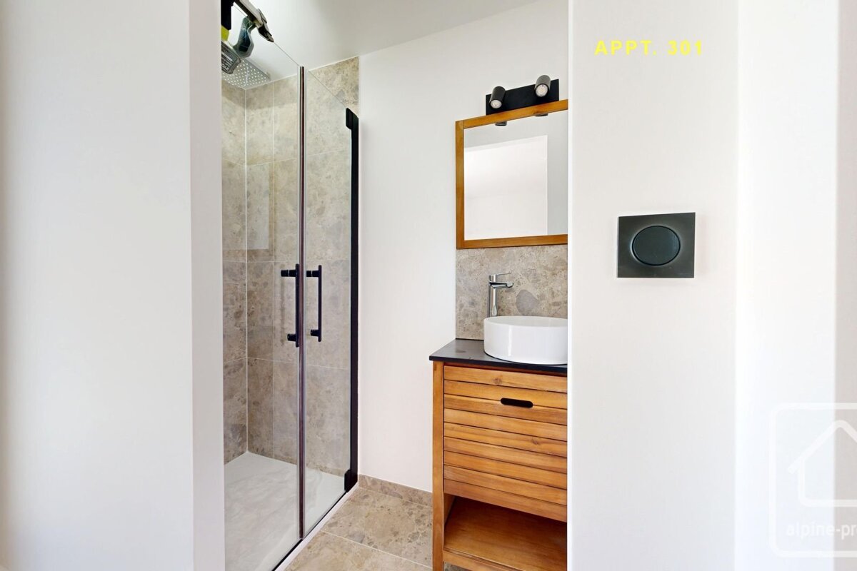 A modern bathroom featuring a glass-enclosed shower, a vanity with a white bowl sink, wooden cabinet, and a mirror with a black light fixture.