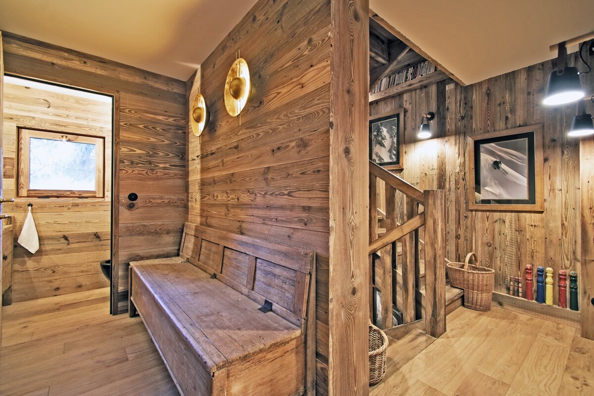 Warm, wood-paneled chalet interior with a rustic bench, staircase, modern lighting, and framed ski art. A doorway leads to another room.