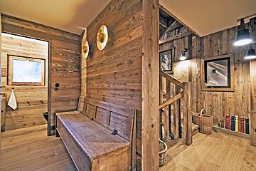 Warm, wood-paneled chalet interior with a rustic bench, staircase, modern lighting, and framed ski art. A doorway leads to another room.