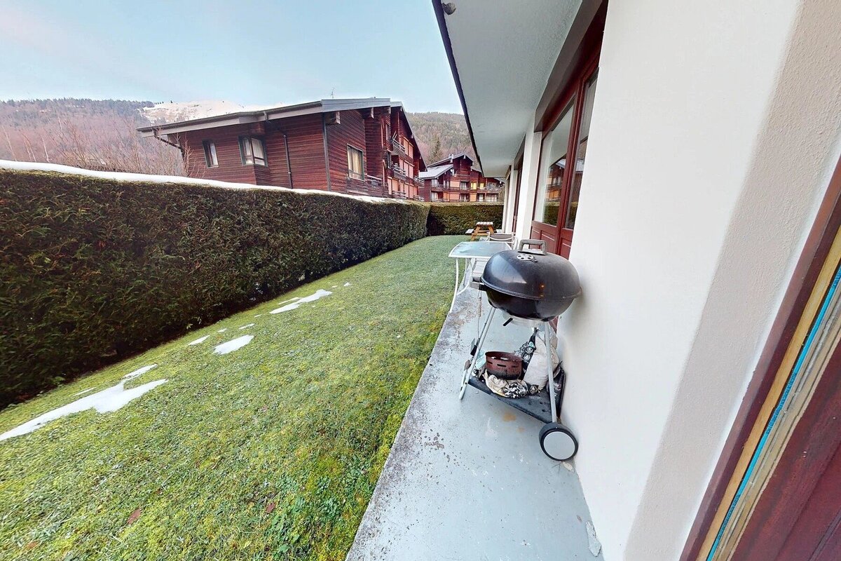 A patio with a grill overlooks a green lawn with snow patches, a tall hedge, and wooden chalets against a mountain backdrop.