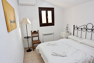 A simple, bright bedroom with white walls, a bed with white linen, a dark-framed window, AC, and a rustic wooden chair.