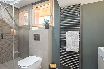 A modern bathroom featuring a glass shower, wall-mounted toilet, heated towel rack, and small window with a plant.