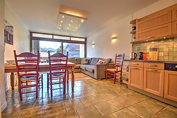 An open-plan living/dining/kitchen area with red chairs, a gray sofa, and wood cabinets. A large window offers mountain views.
