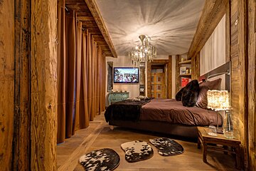 Rustic wooden chalet bedroom with brown curtains, a large bed, cowhide rugs, and an elegant chandelier.