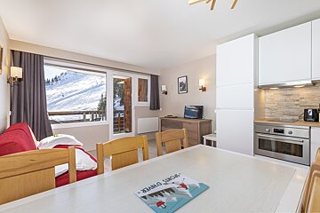 Cozy ski apartment with a red sofa, dining area, modern kitchen, and large windows offering a stunning view of a snowy mountain slope.