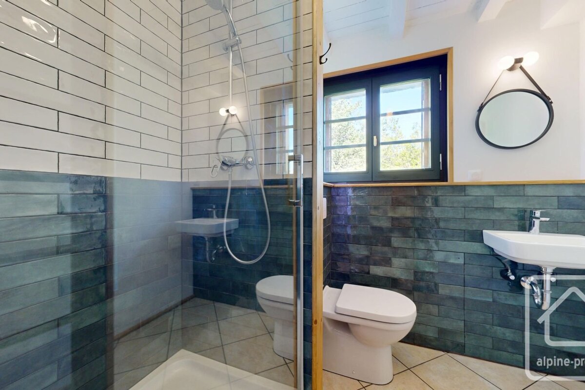 A modern bathroom with blue and white tiled walls, a glass walk-in shower, a toilet, a sink, and a round mirror hanging on the wall.