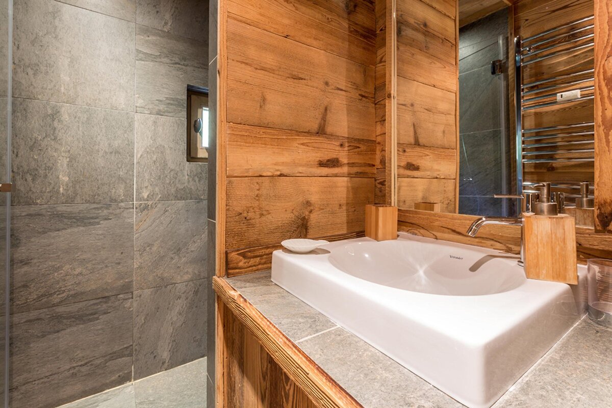 Rustic-modern bathroom with a white sink on a stone counter, warm wood-paneled walls, and a grey slate-tiled shower area.