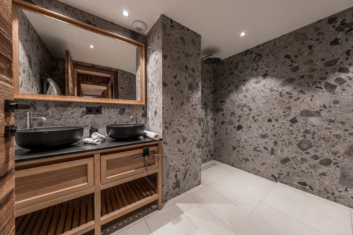 Modern bathroom with a wooden double vanity, black vessel sinks, a large mirror, and grey terrazzo stone walls.