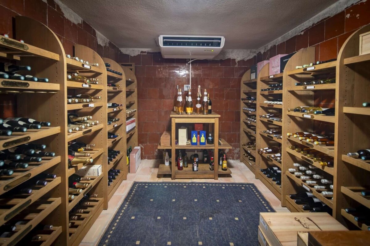A well-stocked wine cellar with wooden racks lining red-tiled walls, a central table, a blue rug, and a cooling unit overhead.